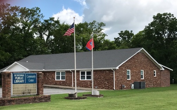 exterior Eagleville Bicentennial Library. One story brick structure with black roof. American flag and Tennessee State flag flying on flag poles. Blue and white sign out front displaying library's name.