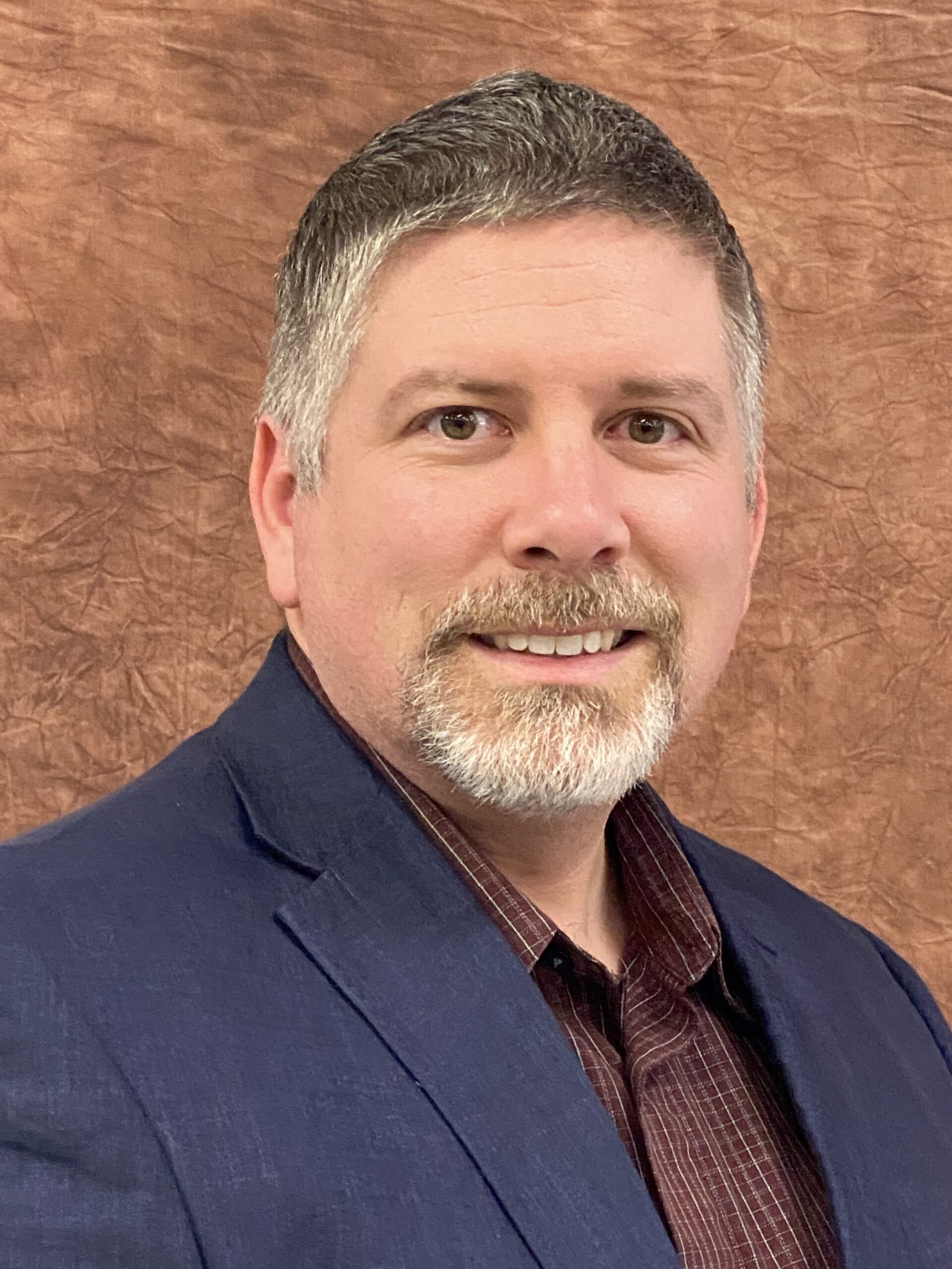 Older white man with greying dark hair and a grey beard and mustache wearing a brown collared button up shirt and navy blue suit jacket