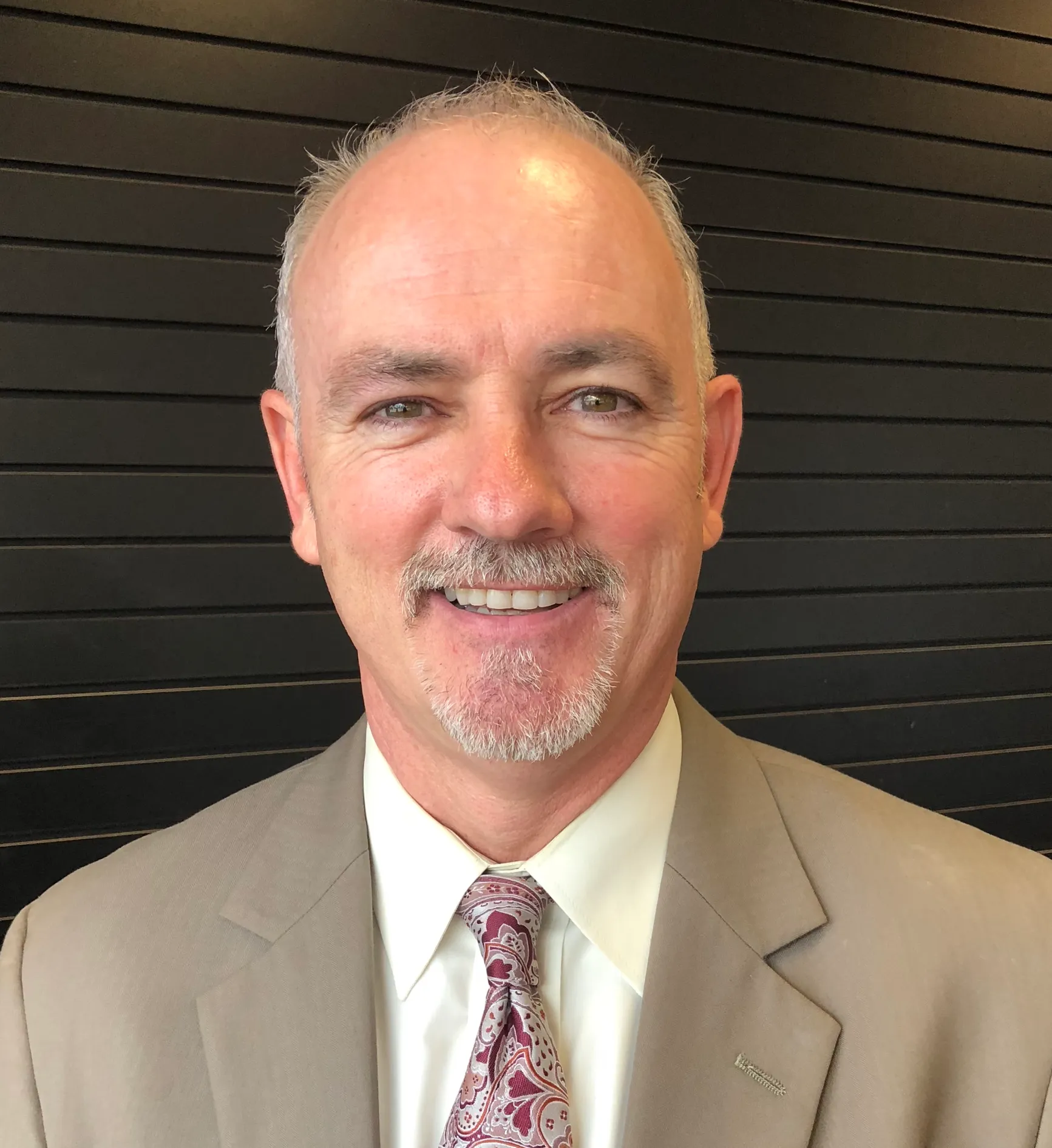 Older white man with thin gray beard and moustache wearing a tan suit jacket and white collared shirt and pink tie