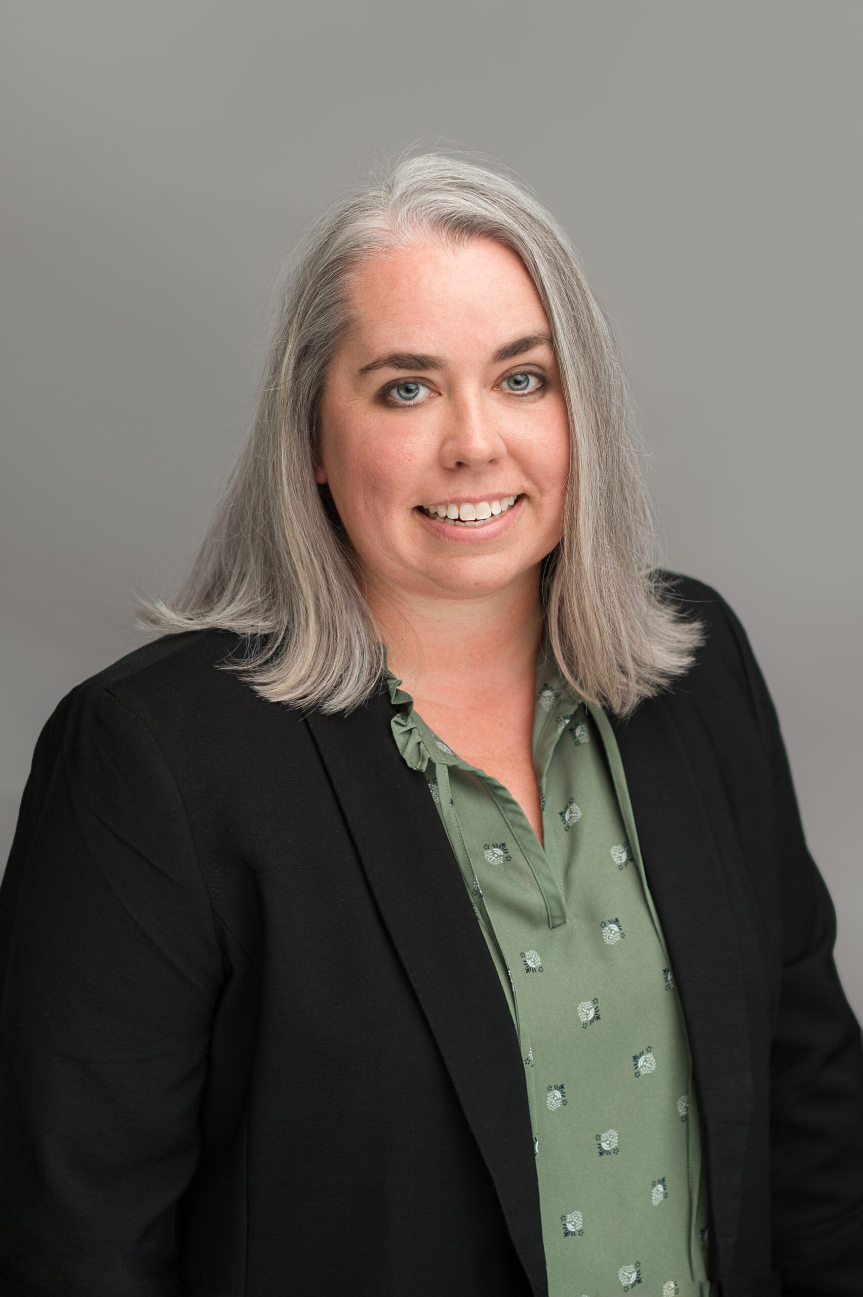 White woman with gray shoulder length hair in a black blazer and green blouse.