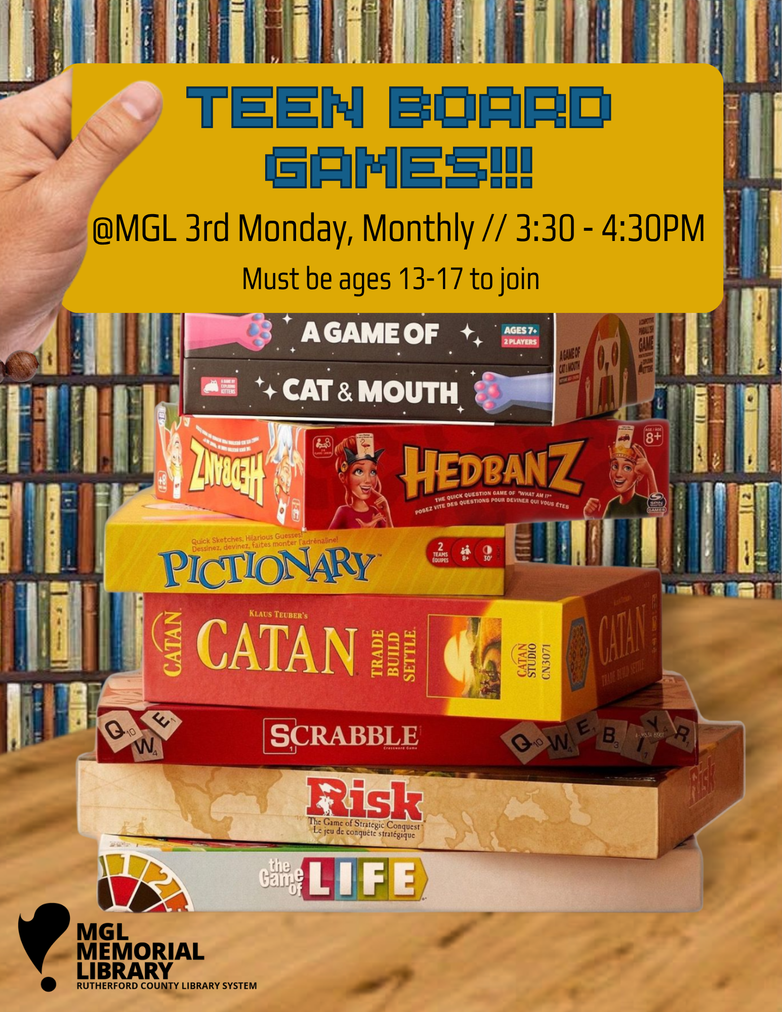stack of common board games such as scrabble and catan and the game of life sitting on a wooden table. library shelves in the background.